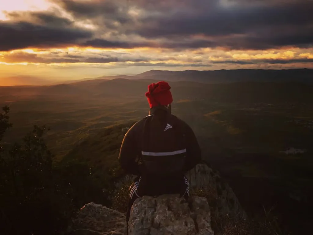Randonneur de dos avec bonnet rouge face à un coucher de soleil sur vallée